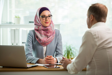 Coworkers meeting in office. Stylish woman in hijab making conversation at desk with man in white...