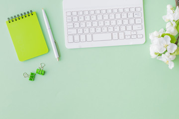 Flat lay blogger or freelancer workspace with a notebook, keyboard and white spring flowers on a green background