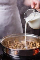 young woman in a gray apron adds milk to mushroom sauce