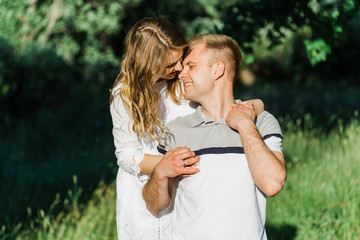 Young pretty couple in love standing in park. Handsome cheerful blonde girl in white dress hugging her boyfriend. Man and woman having fun outdoors