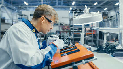 Man in Blue Work Coats is Using Tweezers to Assemble Printed Circuit Boards for Smartphones....