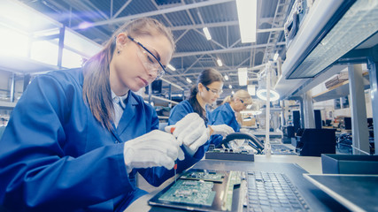 Female Electronics Factory Worker in Blue Work Coat and Protective Glasses is Assembling Laptop's...