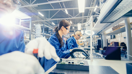 Female Electronics Factory Worker in Blue Work Coat and Protective Glasses is Assembling Laptop's...