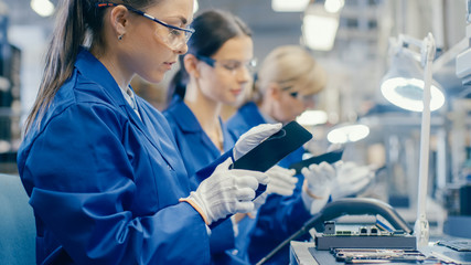 Woman Electronics Factory Worker in Blue Work Coat and Protective Glasses Takes a Smartphone Screen and Performs a Quality Check. High Tech Factory Facility with Multiple Employees. 