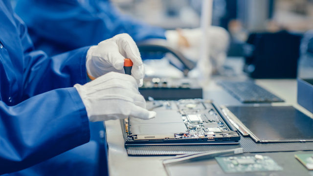 Close-Up of a Female Electronics Factory Worker in Blue Work Coat Assembling Laptop's Motherboard with a Screwdriver. High Tech Factory Facility with Multiple Employees. 