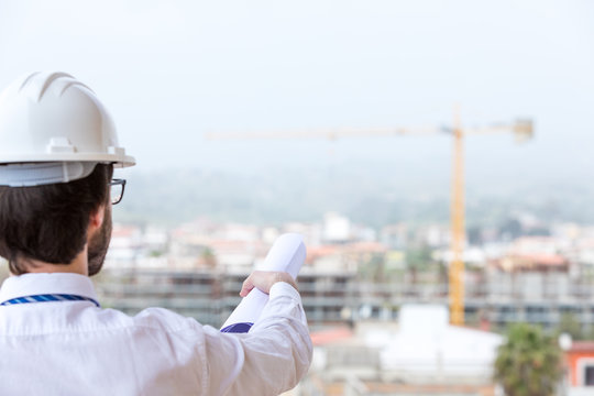 Giovane ingegnere edile con occhiali da vista, caschetto e camicia bianco, cravatta blu. Di fronte a un cantiere edile con gru e palazzina in costruzione visto di spalle.