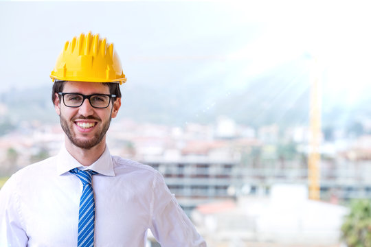 Giovanissimo architetto edile con caschetto giallo, occhiali da vista, camicia bianca e cravatta blu sorride in primo piano con sfondo del cantiere sfuocato.