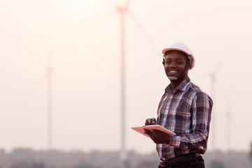 African engineer wearing white hard hat standing with digital tablet against wind turbine on sunny day