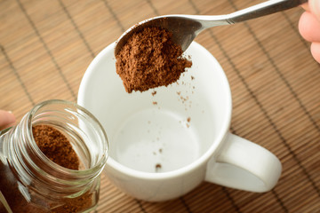 Hand pours ground coffee from a spoon in a coffee cup. Holding jar with coffee in the other hand