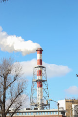 Red and white stripe power plant tube with white smoke over blue sky. rowns of trees without leaves. Air pollution, industrial emissions.