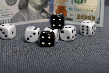 White dice with black markings. They lie on a surface covered with a coarse gray cloth. Among them are two black dice with white markings. Nearby is a hundred dollar bill.