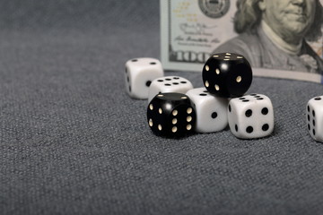 White dice with black markings. They lie on a surface covered with a coarse gray cloth. Among them are two black dice with white markings. Nearby is a hundred dollar bill.