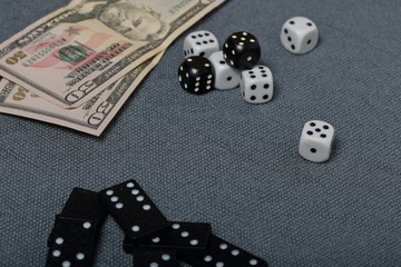 White dice with black markings. They lie on a surface covered with a coarse gray cloth. Among them are two black dice with white markings. Next door are dominoes and dollars.