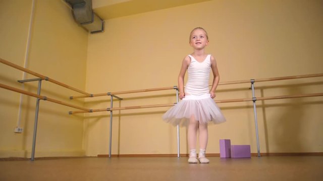 A little blonde girl in a white swimsuit puts on a tutu skirt in a ballet studio.