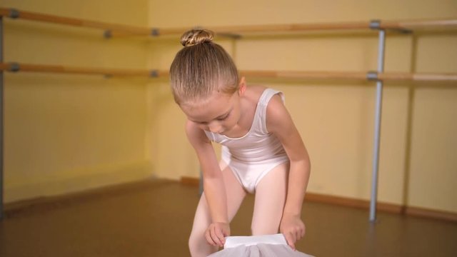 A little blonde girl in a white swimsuit puts on a tutu skirt in a ballet studio.