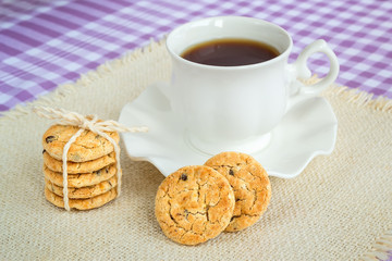 Stack of oatmeal cookies with pieces of dark chocolate and porcelain cup with tea on saucer with wavy edge. Sweet breakfast.