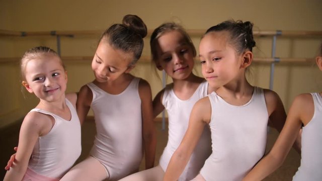 Little ballerina girls in white swimsuits look directly into the camera during classes in the ballet studio.