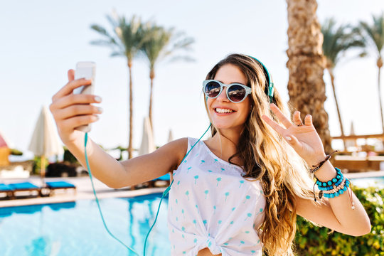 Happy girl in sunglasses with tanned skin making selfie with peace sign on palm trees background. Charming long-haired woman taking photo of herself near the pool while resting on resort in sunny day