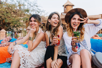 Three pretty female tourists in trendy outfit drink cocktails while resting after sightseeing. Laughing long-haired girls having fun in outdoor cafe, sitting on colorful sofa and waiting for boys