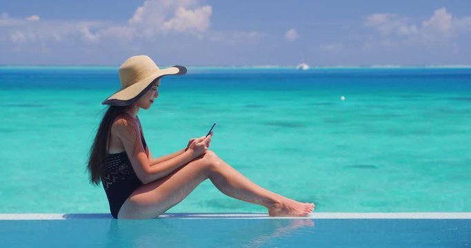 Phone - woman using mobile cell phone app on vacation sitting af pool by tropical ocean coral lagoon on travel holidays. Slow motion shot on RED.