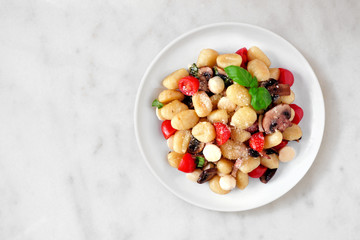 Gnocchi with tomatoes, mozzarella, mushrooms and basil. Top view over a white marble background.