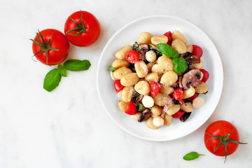 Gnocchi with tomatoes, mozzarella, mushrooms and basil. Above view scene over a white marble background.