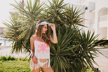 Attractive girl in trendy hat and vintage tank-top with fringe looking away, covering eyes from sunlight. Pretty young woman in stylish shirt and white panties standing near the palm tree