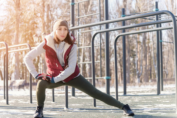 Sports girl in warm clothes on a sunny day does a warm-up on the outdoor sports field in the winter...