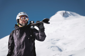 Fototapeta na wymiar Portrait bearded male skier aged against background of snow-capped Caucasus mountains. An adult man wearing ski googles mask and helmet skis on his shoulder looks mountains. Ski resort concept