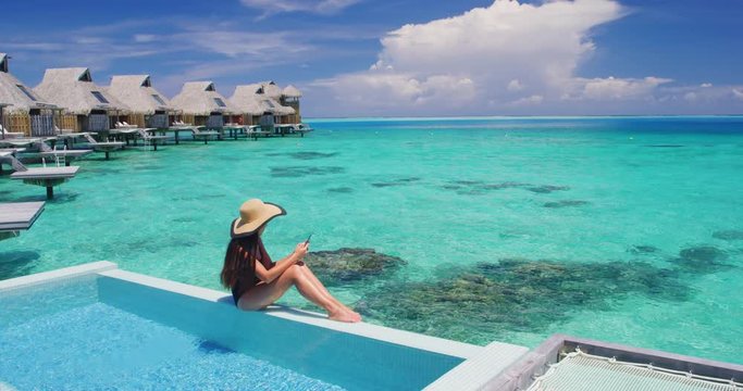 Woman using mobile cell phone app on vacation sitting af pool by tropical ocean coral lagoon on travel holidays. Slow motion.