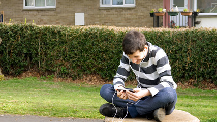 teen boy with smart phone listening or talking while sitting on stump in british park. teenager and social media concept