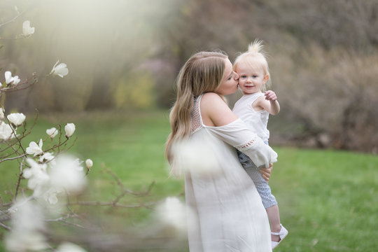 Beautiful young mother kissing toddler daughter on cheek in spring blossoms