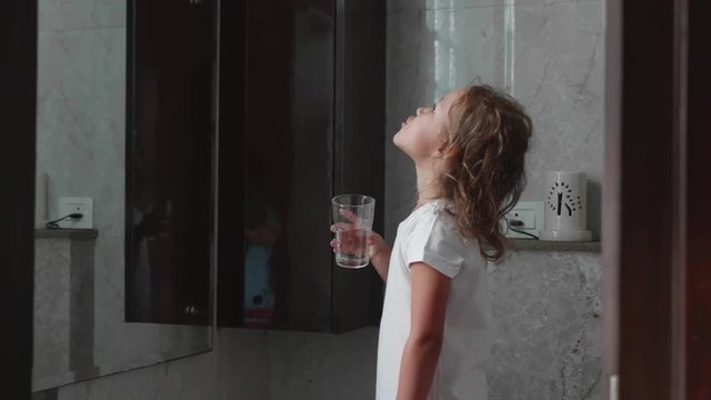 Cute little curly child girl rinses her mouth with water, looking at mirror and spits water into the sink, at domestic bathroom, side view.