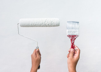 Woman holding up used paint brush and roller with white paint and white wall background.