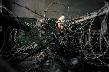 Young girl is sitting behind barbwire in old, ruined factory. - 264701253