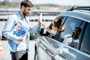 Instructor giving a five to a woman driver sitting in the car while learning to drive on the training ground