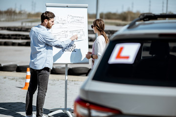 Male instructor showing traffic shemes to a young female student standing on the training ground at the driver's school