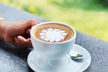 Hands are holding a cup of hot latte coffee on a natural blurred background.
