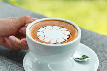 Hands are holding a cup of hot latte coffee on a natural blurred background.