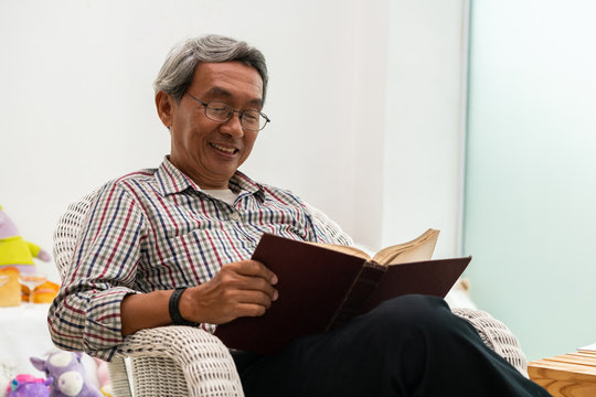 Happy Senior Asian Man Reading Book On The Chair In Living Room At Home. Retirement Lifestyle And Aging Society Concept.