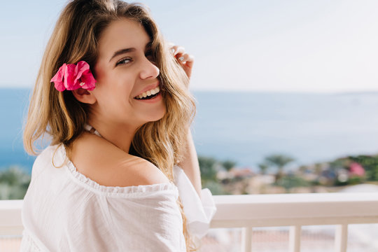 Cheerful laughing girl with cute flower in light-brown hair posing on horizon background. Gorgeous young woman in white outfit enjoying her vacation on resort and spending time on fresh air