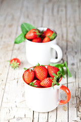 Organic red strawberries in two white ceramic cups and mint leaves on rustic wooden background.