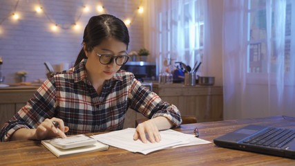 Fototapeta na wymiar Portrait of young asian woman sitting at home kitchen table at night filling application form calculating expenses trying to solve budget problems looking at papers managing with utility bills.