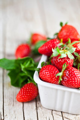 Fresh red strawberries in white bowl and mint leaves on rustic wooden background.
