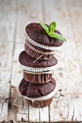 Three fresh dark chocolate muffins with mint leaves on rustic wooden table.