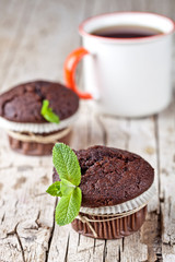 Fresh dark chocolate muffins with mint leaves and cup of tea on rustic wooden table.