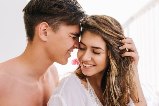 Dreamy girl with excited smile holding her hair and listening love confession from handsome young man with dark short hair. Portrait of boy and his girlfriend spending time together in morning