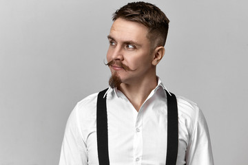 Style, fashionand elegance concept. Horizontal shot of handsome elegant man with stylish mustache and goatee posing at blank gray wall, wearing suspenders over white shirt, looking away with smile