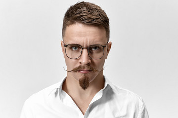 Horizontal shot of displeased annoyed businessman in glasses and formal white shirt posing at blank studio wall, staring at camera with angry strict look, mad at his secretary because of cold coffee