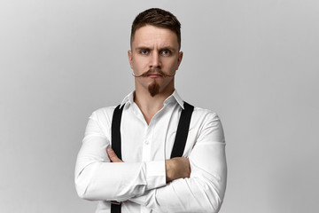 Stylish confident bearded boss in suspenders and white shirt posing isolated at blank copyspace studio wall, crossing arms on his chest, angry with inefficient business strategy, having strict look